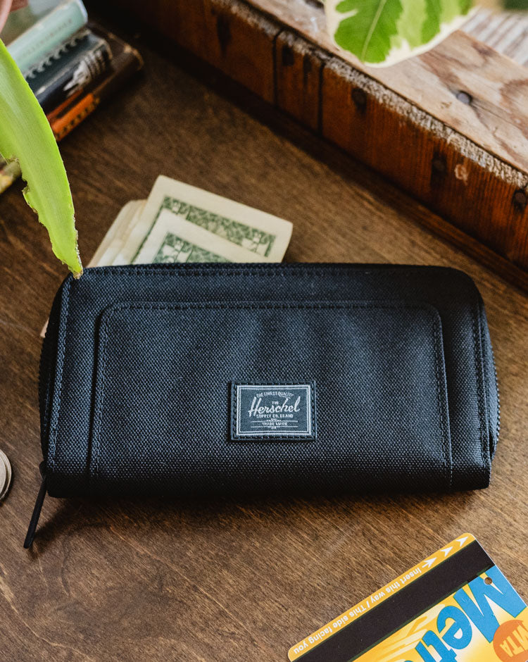 Front view of the Herschel Thomas Wallet in Black Tonal, showing its sleek, zippered accordion-style design and signature woven label.