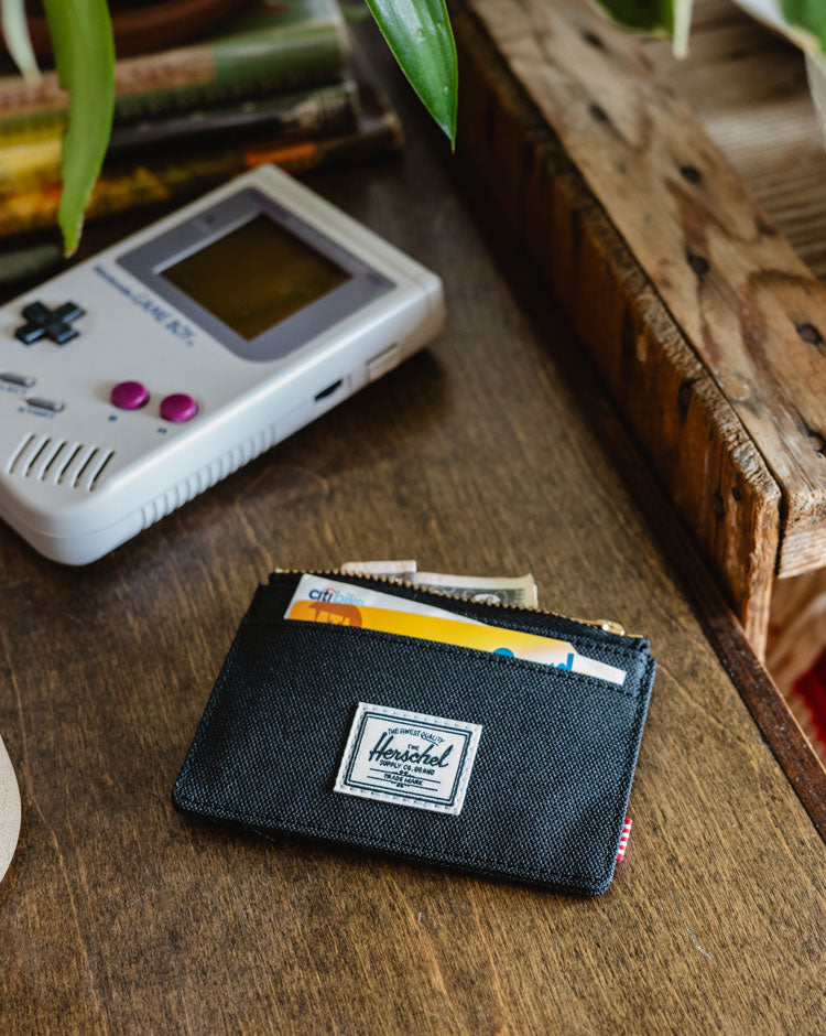 Black Herschel Supply Co. Oscar Cardholder shown in a lifestyle mobile view, featuring multiple card slots and the signature woven label.
